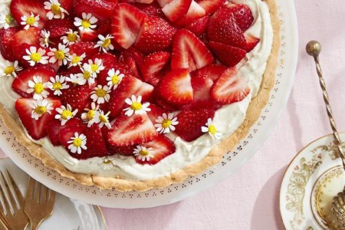 A tart topped with sliced strawberries and small white flowers on a white plate, surrounded by tea cups, saucers, and gold utensils-a delightful choice for Memorial Day Desserts on a pink tablecloth.