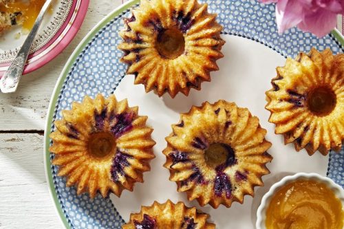 Six sunflower-shaped muffins with berry centers are arranged on a blue-patterned plate, accompanied by a small bowl of jam and a fork, making them perfect Memorial Day Desserts on a striped cloth beside flowers.