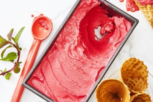 Rectangular pan with bright pink sorbet, a pink ice cream scoop, waffle cones, a scoop of sorbet on a cone, and mint sprigs on a white surface-perfect for colorful Memorial Day Desserts.