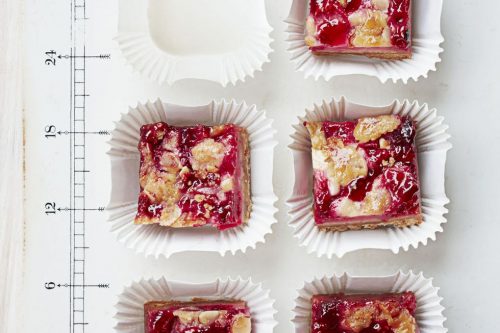 Six square fruit crumble bars are arranged in white paper cups on a white board with measurement markings, with one empty cup in the top row-perfect as Memorial Day Desserts for your summer celebration.