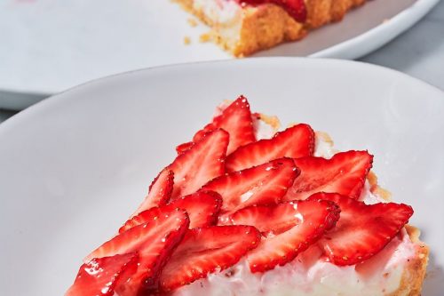 A slice of strawberry tart with a creamy filling and crumbly crust is served on a white plate, making it a perfect choice for Memorial Day Desserts, with the rest of the tart in the background.