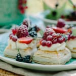 Puff pastry tarts are served as delicate squares topped with whipped cream, raspberries, red currants, blackberries, and sliced strawberries on a plate.