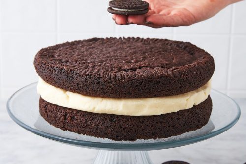 A hand places a chocolate sandwich cookie on top of a large chocolate cake with cream filling, displayed on a glass cake stand. Perfect for Father's Day Dessert Recipes, more cookies are scattered on the marble surface below.