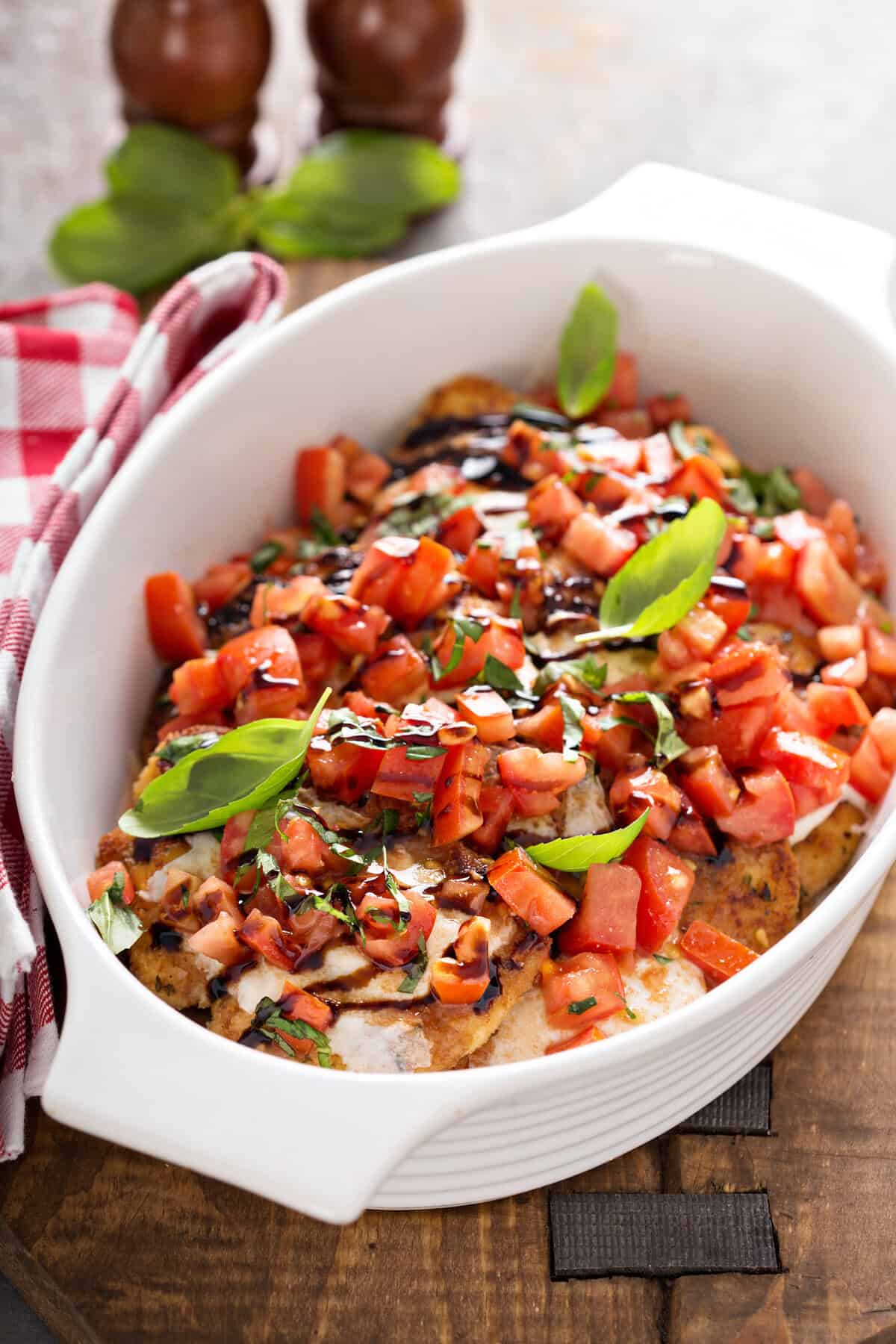 A white baking dish filled with bruschetta chicken—juicy chicken breast topped with chopped tomatoes, fresh basil leaves, and a drizzle of balsamic glaze—is placed on a wooden board beside a red checkered towel.