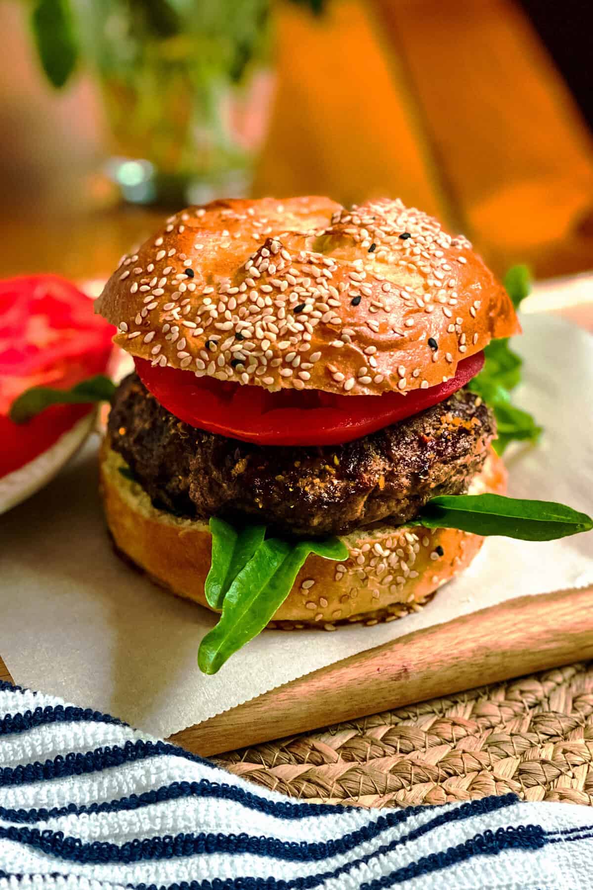 A close-up of a sesame seed burger bun filled with a grilled beef patty, fresh tomato slices, and green leafy spinach—perfect for trying out new Weber Seasoning recipes—rests on parchment paper atop a wooden board. A striped cloth is visible in the foreground.