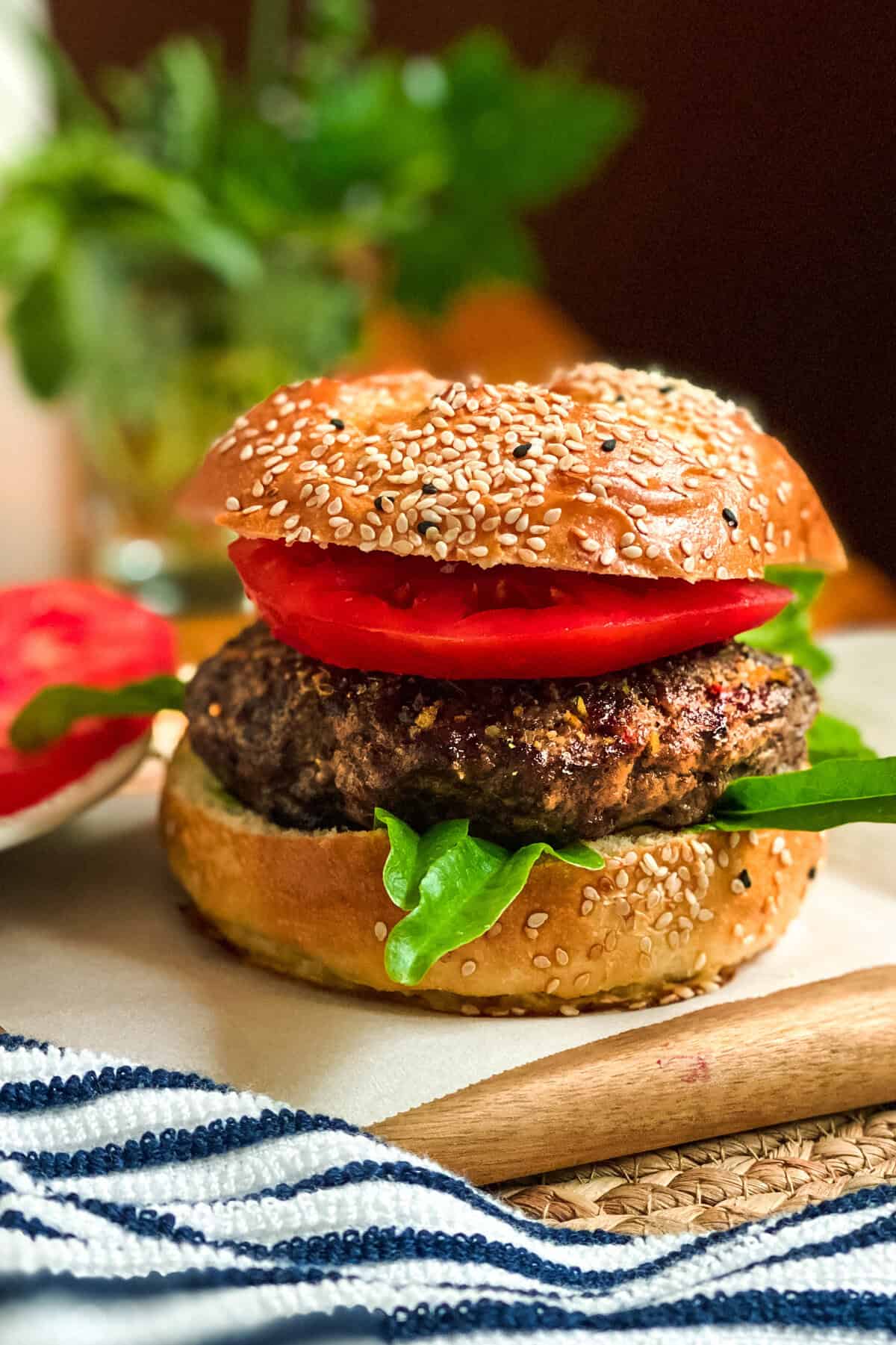 A close-up of a sesame seed bun burger with leafy greens, a thick beef patty, and two slices of tomato sits on parchment paper-perfect inspiration for your next burgers recipe-striped cloth and blurred greenery in the background.