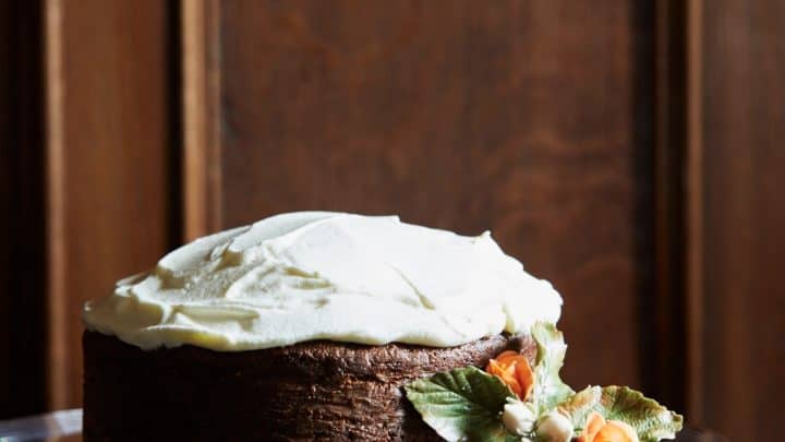 Chocolate cake with white frosting sits on a glass cake stand, decorated with small orange flowers and green leaves, reminiscent of elegant Queen Elizabeth Afternoon Tea Recipes, in front of a wooden background.