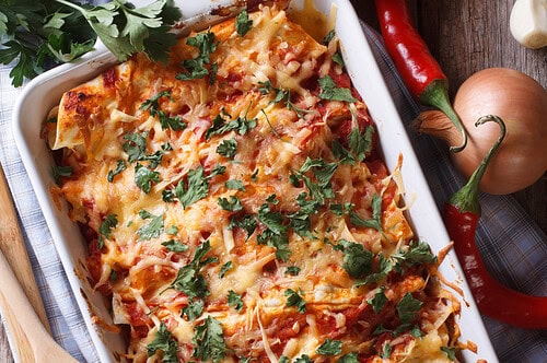 A baked dish of leftover chicken enchiladas topped with melted cheese and chopped cilantro sits on a rustic wooden table, surrounded by fresh tomatoes, parsley, red chili peppers, garlic, onion, and wooden utensils.