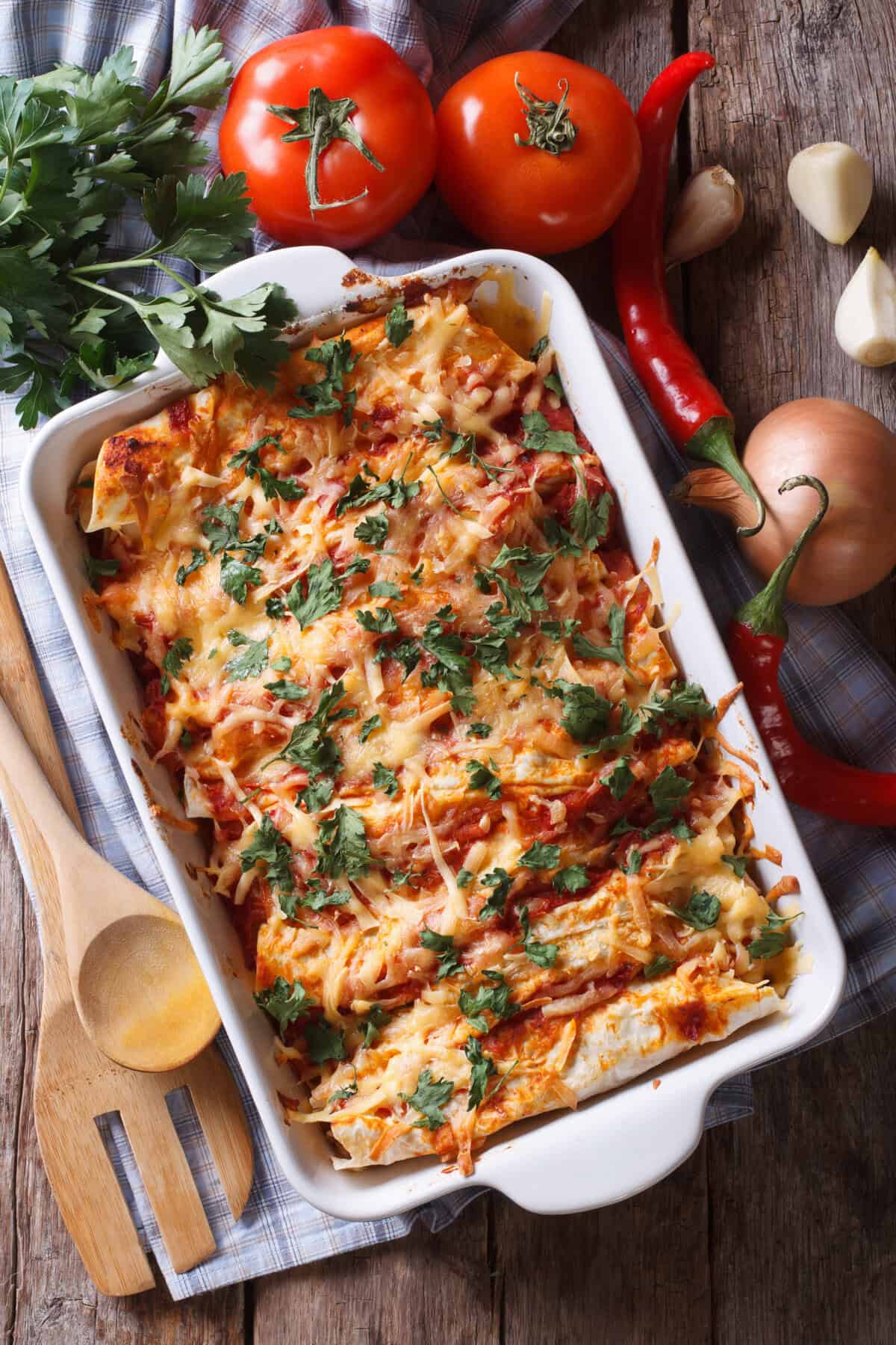 A baked dish of leftover chicken enchiladas topped with melted cheese and chopped cilantro sits on a rustic wooden table, surrounded by fresh tomatoes, parsley, red chili peppers, garlic, onion, and wooden utensils.