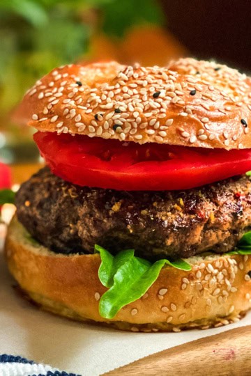 Grilled beef burger with tomatoes and lettuce on a sesame bun with tomato slices nearby.