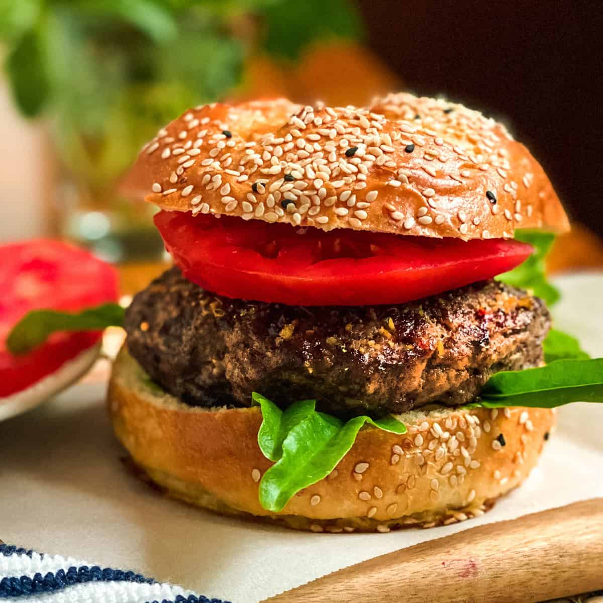 Grilled beef burger with tomatoes and lettuce on a sesame bun with tomato slices nearby.