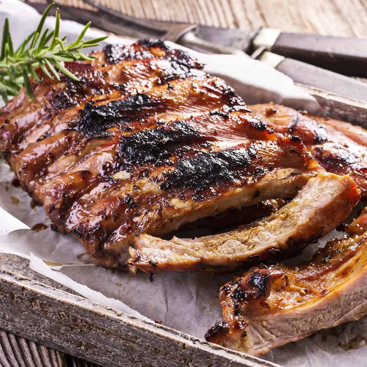 A rack of grilled barbecue ribs with a slightly charred surface, perfect for a 4th of July Cookout, garnished with rosemary and served on parchment paper in a rustic wooden tray.