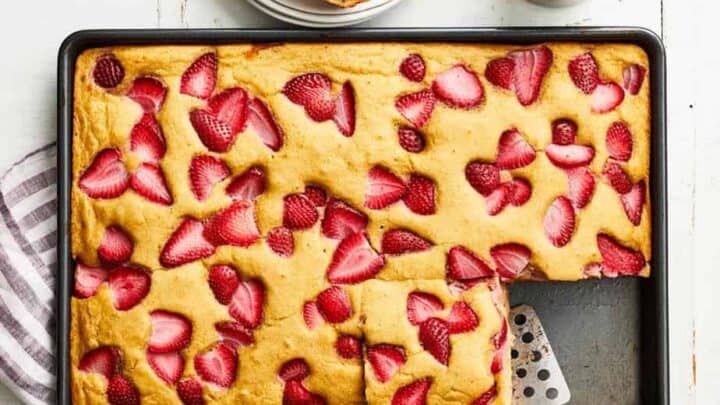 A rectangular baking pan with strawberry sheet cake—perfect for a sheet pan breakfast—one square piece removed and placed on a spatula, next to a plate with two pieces and a small pitcher of syrup.