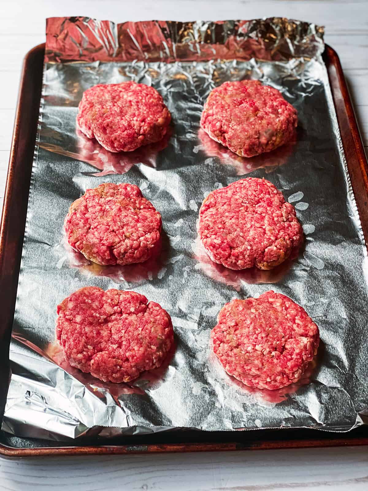 Six raw beef burger patties arranged on a foil-lined baking sheet, ready to be cooked for your next burgers recipe. The tray is set on a light-colored surface.