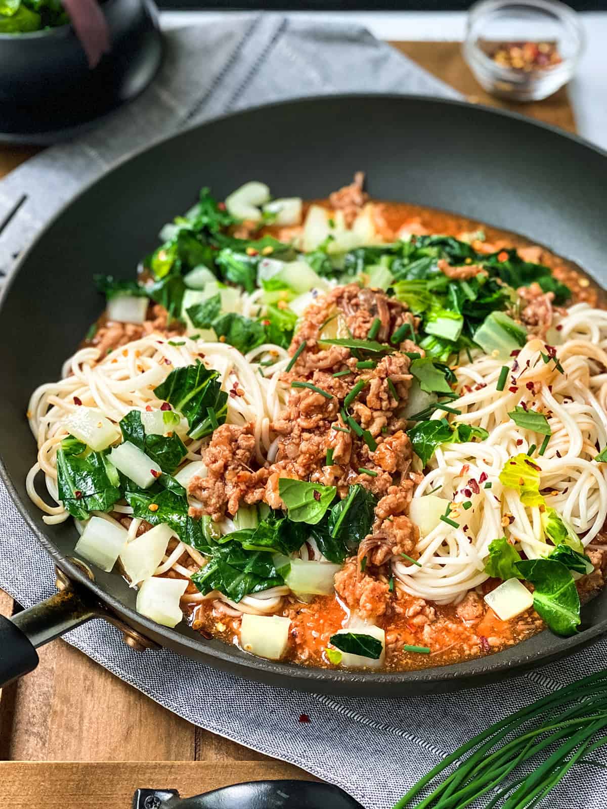 A pan with cooked ginger pork noodles, ground meat, chopped bok choy, and a red sauce, topped with sliced green onions and red pepper flakes.