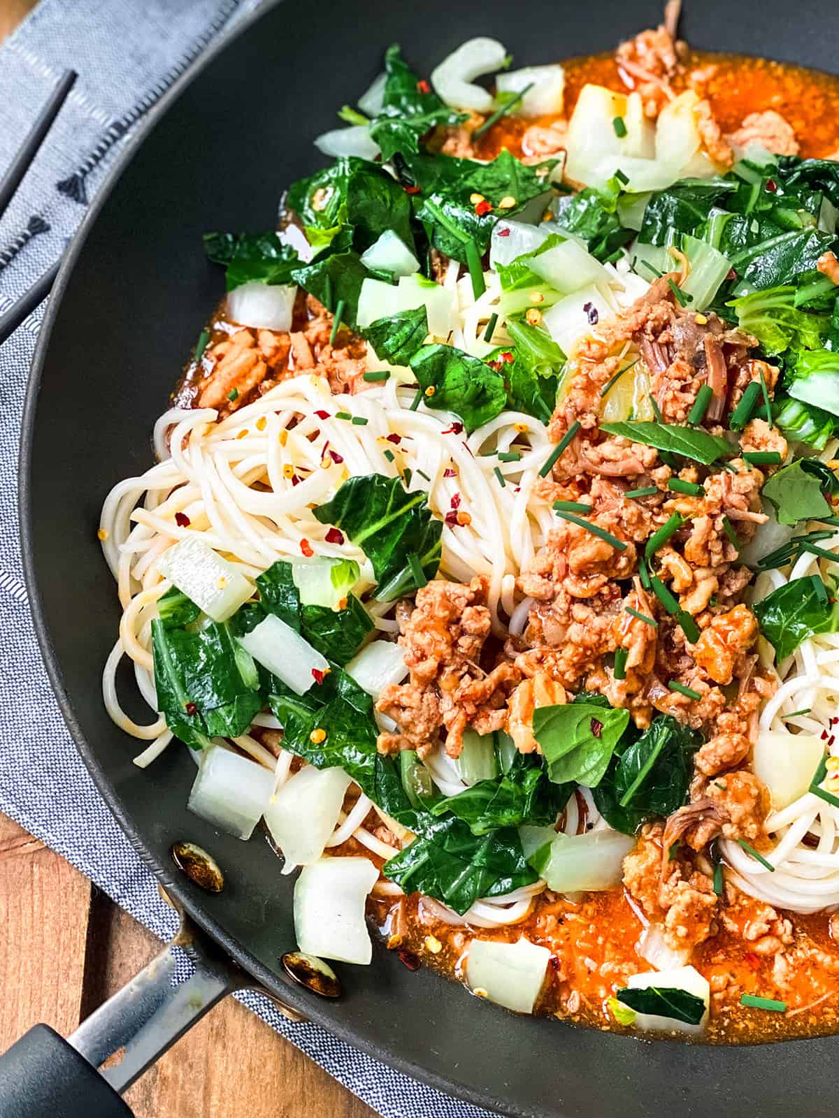 A skillet with cooked ginger pork noodles, ground meat, chopped bok choy, leafy greens, and red sauce, placed on a blue cloth on a wooden table.