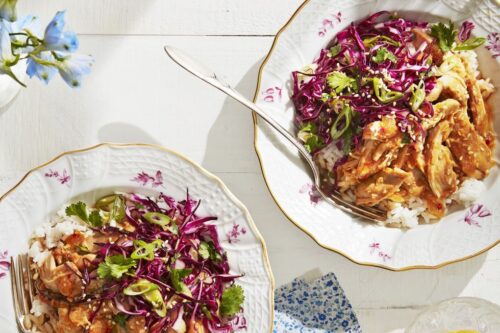 Two plates of Summer Slow Cooker shredded chicken over rice with purple cabbage slaw and cilantro, set on a white table with glasses of water and a blue patterned napkin.