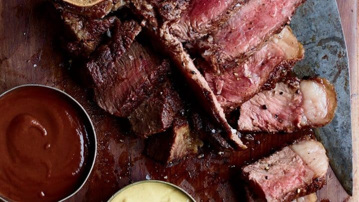 Closeup top view of cooked porterhouse steak with BBQ sauce and horseradish sauce on a wooden cutting board.