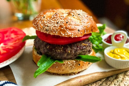 A sesame seed bun burger with a thick beef patty, fresh tomato slices, and leafy greens, served on parchment paper with sides of ketchup and mustard-perfect inspiration for your next burgers recipe.