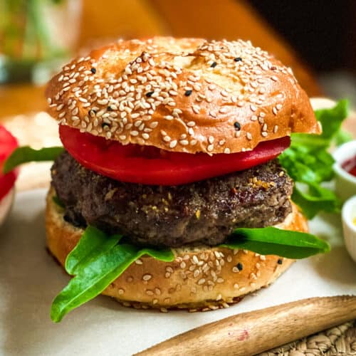 A sesame seed bun burger with a thick beef patty, fresh tomato slices, and leafy greens, served on parchment paper with sides of ketchup and mustard—perfect inspiration for your next burgers recipe.