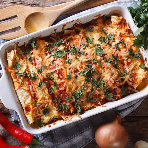 A baking dish of cheesy leftover chicken enchiladas topped with fresh herbs sits on a checkered cloth, surrounded by tomatoes, parsley, a red chili pepper, an onion, garlic cloves, and wooden utensils on a wooden table.