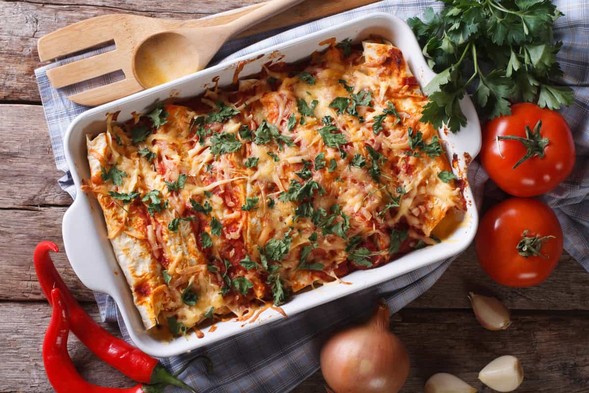 A baking dish of cheesy leftover chicken enchiladas topped with fresh herbs sits on a checkered cloth, surrounded by tomatoes, parsley, a red chili pepper, an onion, garlic cloves, and wooden utensils on a wooden table.