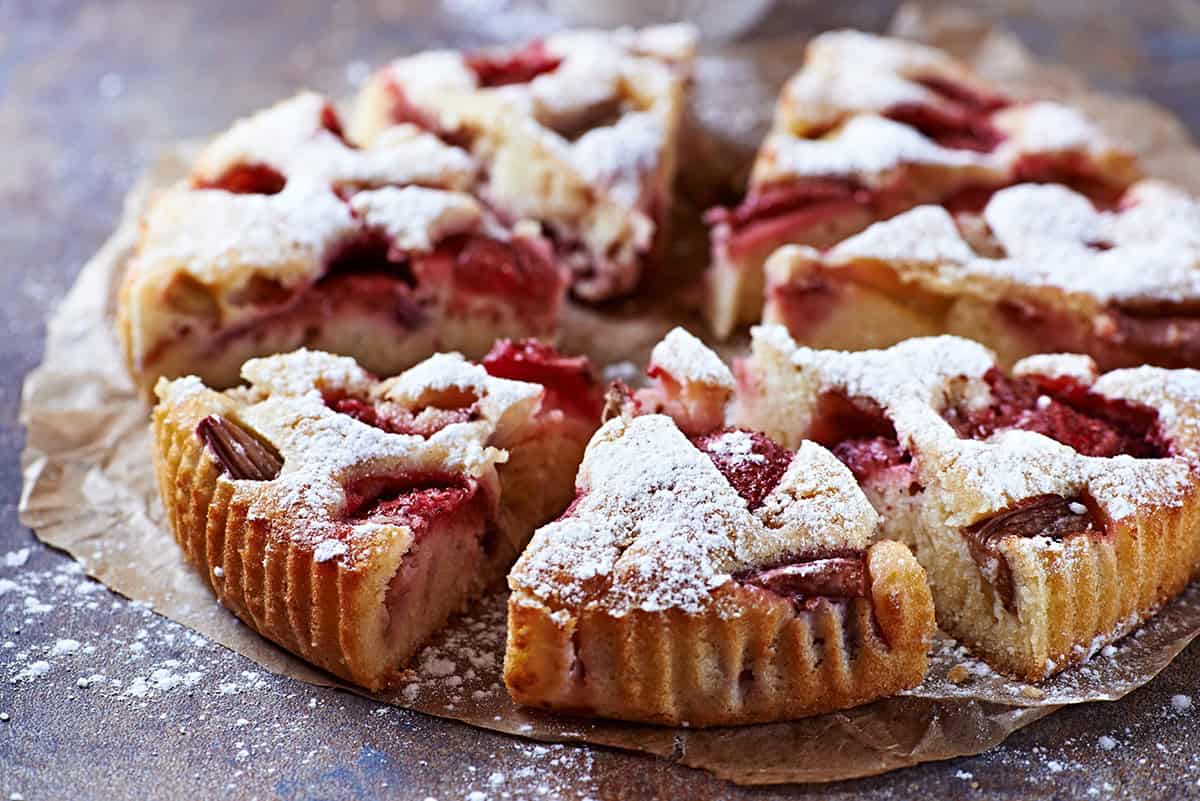 Sliced round strawberry rhubarb cake with visible strawberries and almonds, dusted with powdered sugar, arranged on parchment paper.