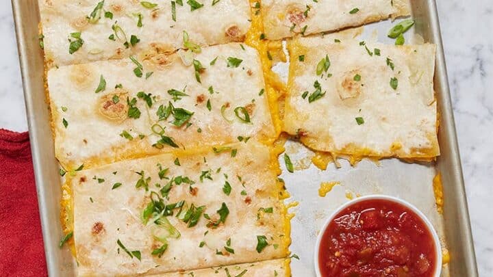 A baking sheet with rectangular quesadilla slices topped with chopped herbs, a bowl of salsa, and a small dish of sour cream on the side—perfect for those seeking healthy lunch ideas.