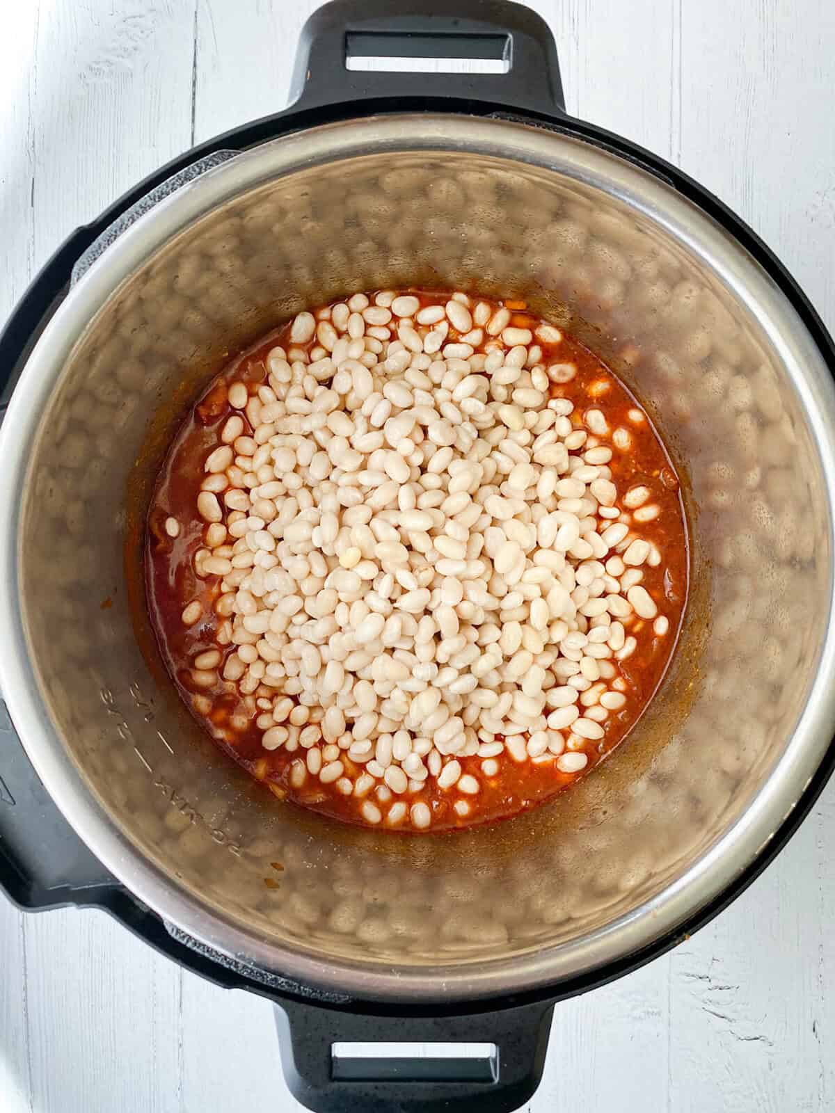 A top view of an Instant Pot filled with white beans and red sauce, ready to become delicious Instant Pot Baked Beans. The pot sits on a white wooden surface, prepared for cooking.