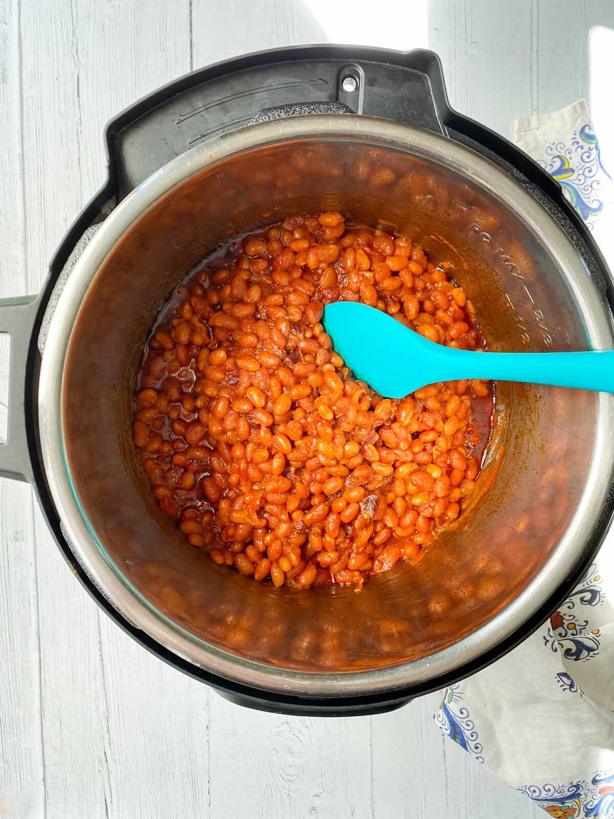 A pot of Instant Pot Baked Beans in a tomato-based sauce sits inside an Instant Pot, with a bright blue spatula resting in the beans. The scene is lit by natural sunlight on a white wooden surface.