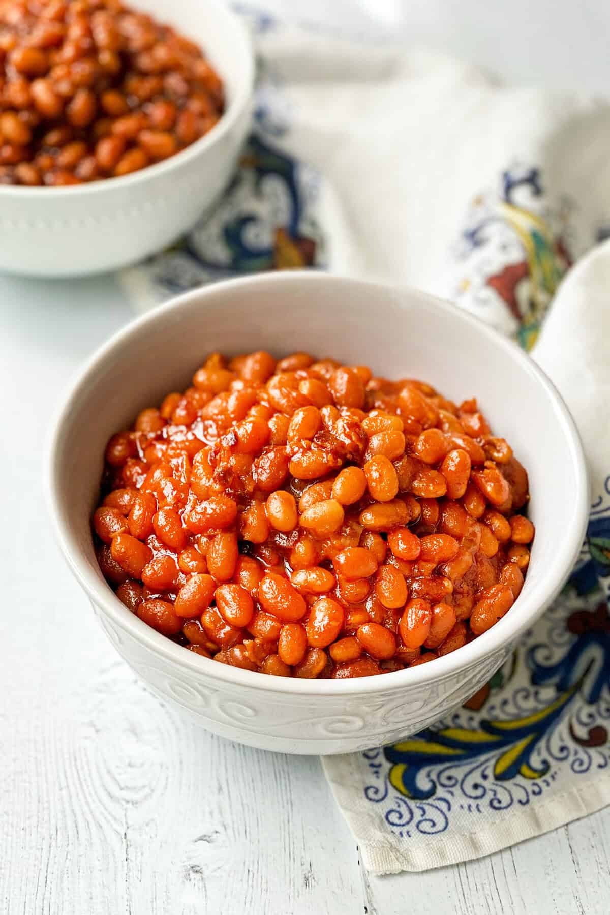A white bowl filled with Instant Pot Baked Beans in a rich tomato sauce sits on a patterned cloth, with another bowl of beans blurred in the background on a white wooden surface.
