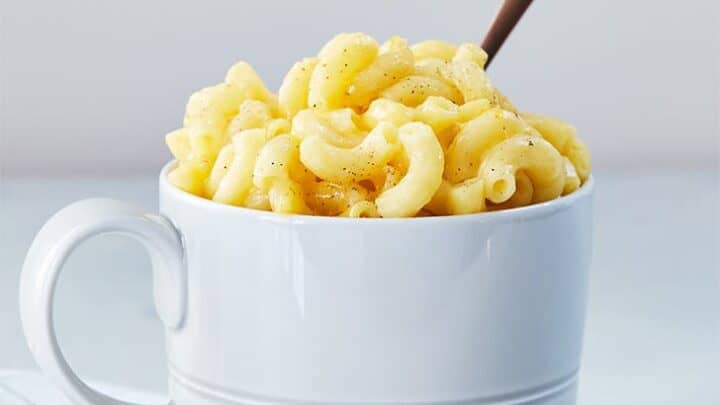A white mug filled with macaroni and cheese, topped with black pepper, sits on a folded white and blue checkered cloth with a spoon inside—perfect for those seeking quick and tasty healthy lunch ideas.