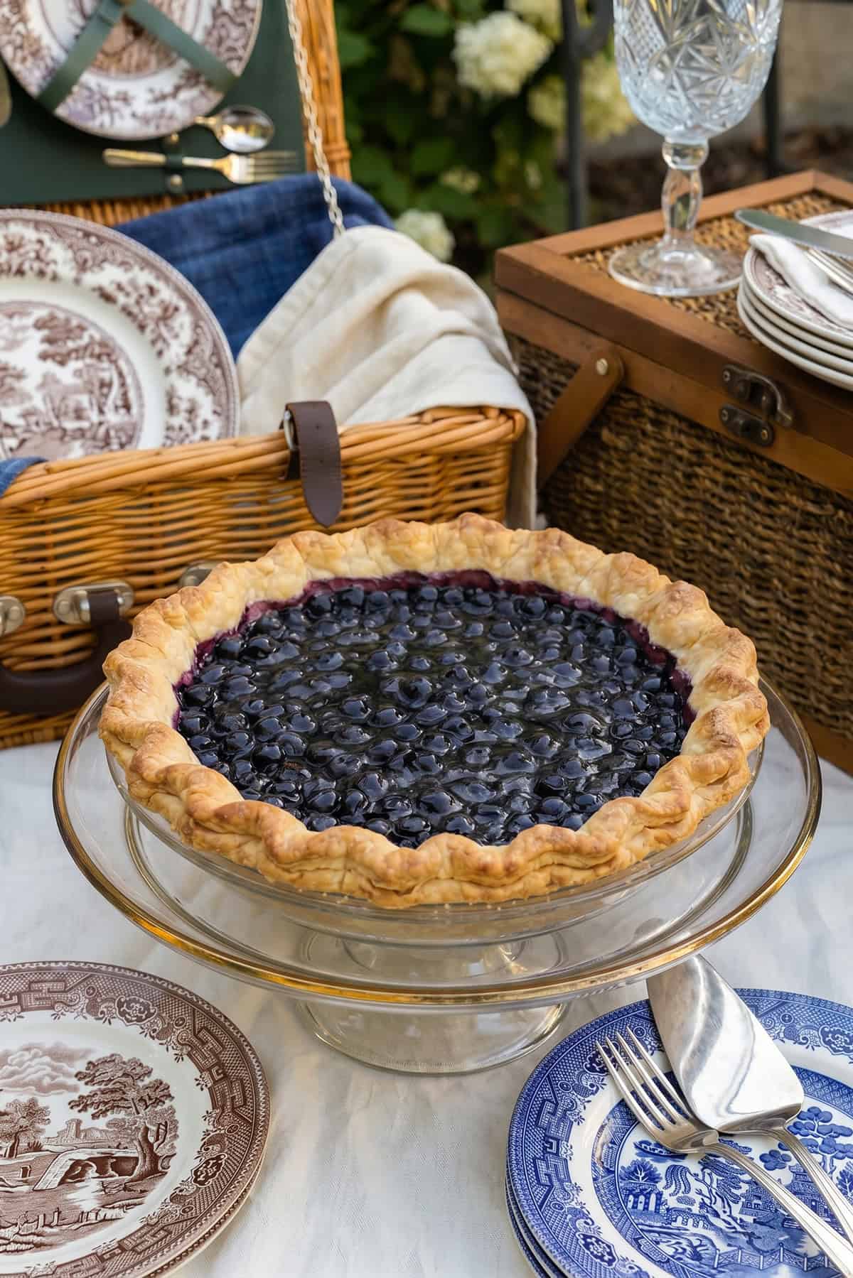 A freshly baked open face blueberry pie on a glass stand sits on a picnic table, surrounded by plates, utensils, and wicker baskets in the background.
