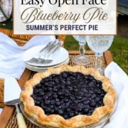 A homemade blueberry pie with a golden crust sits on a wicker table next to two empty plates, silverware, and glasses, with a summer garden in the background.