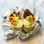 A burrito cut in half reveals rice, bulgogi shredded beef, lettuce, carrots, and other vegetables, placed on foil on a marble surface.