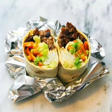 A burrito cut in half reveals rice, bulgogi shredded beef, lettuce, carrots, and other vegetables, placed on foil on a marble surface.