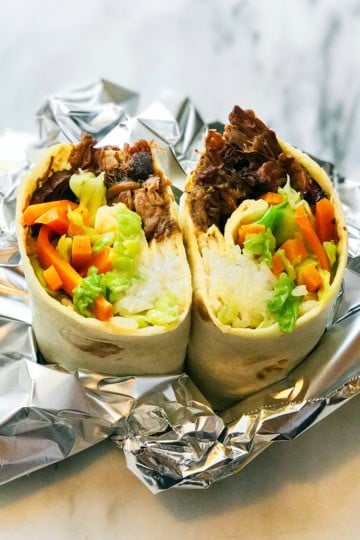 A burrito cut in half reveals rice, bulgogi shredded beef, lettuce, carrots, and other vegetables, placed on foil on a marble surface.