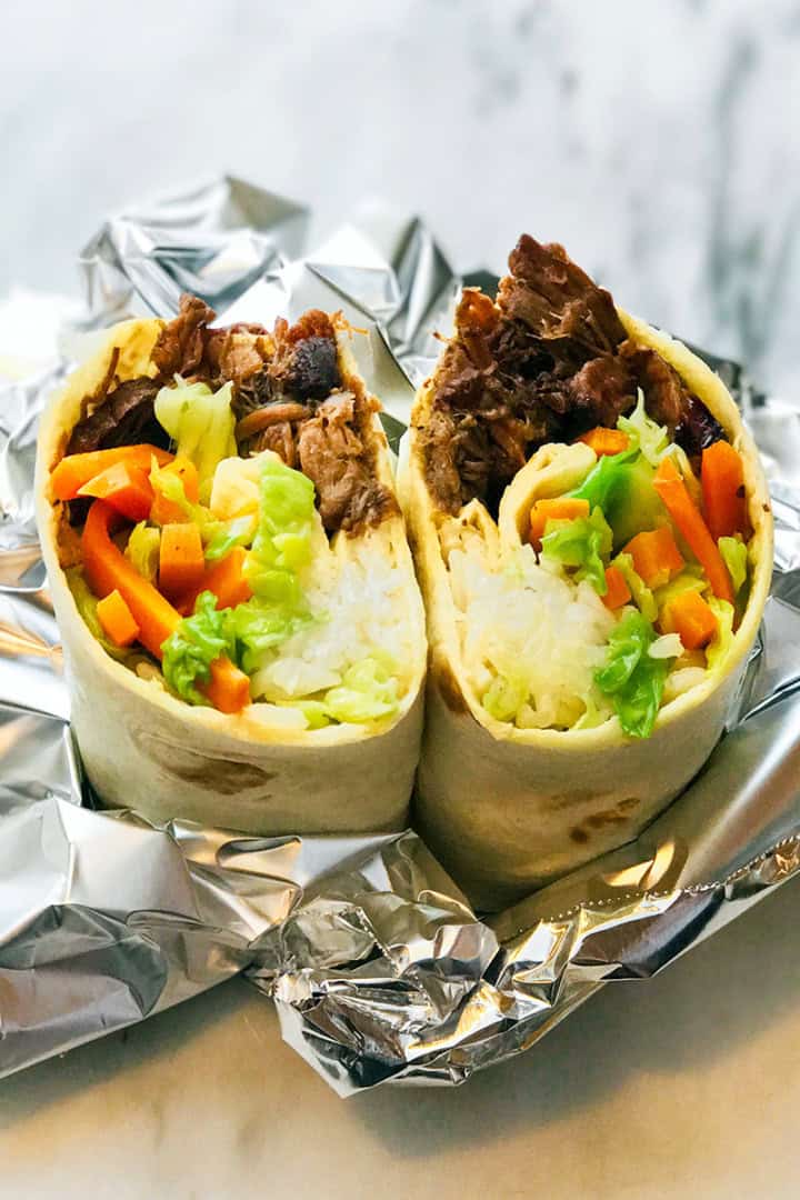 A burrito cut in half reveals rice, bulgogi shredded beef, lettuce, carrots, and other vegetables, placed on foil on a marble surface.