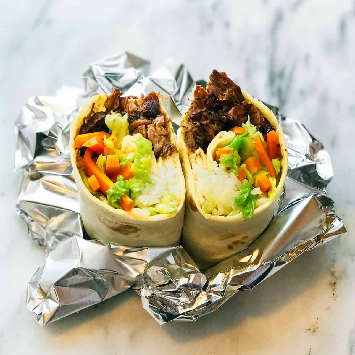 A burrito cut in half reveals rice, bulgogi shredded beef, lettuce, carrots, and other vegetables, placed on foil on a marble surface.