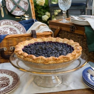 A freshly baked open face blueberry pie sits on a glass cake stand, surrounded by patterned plates, cutlery, and a wicker picnic basket on an outdoor table, ready for a delightful blueberry pie picnic.