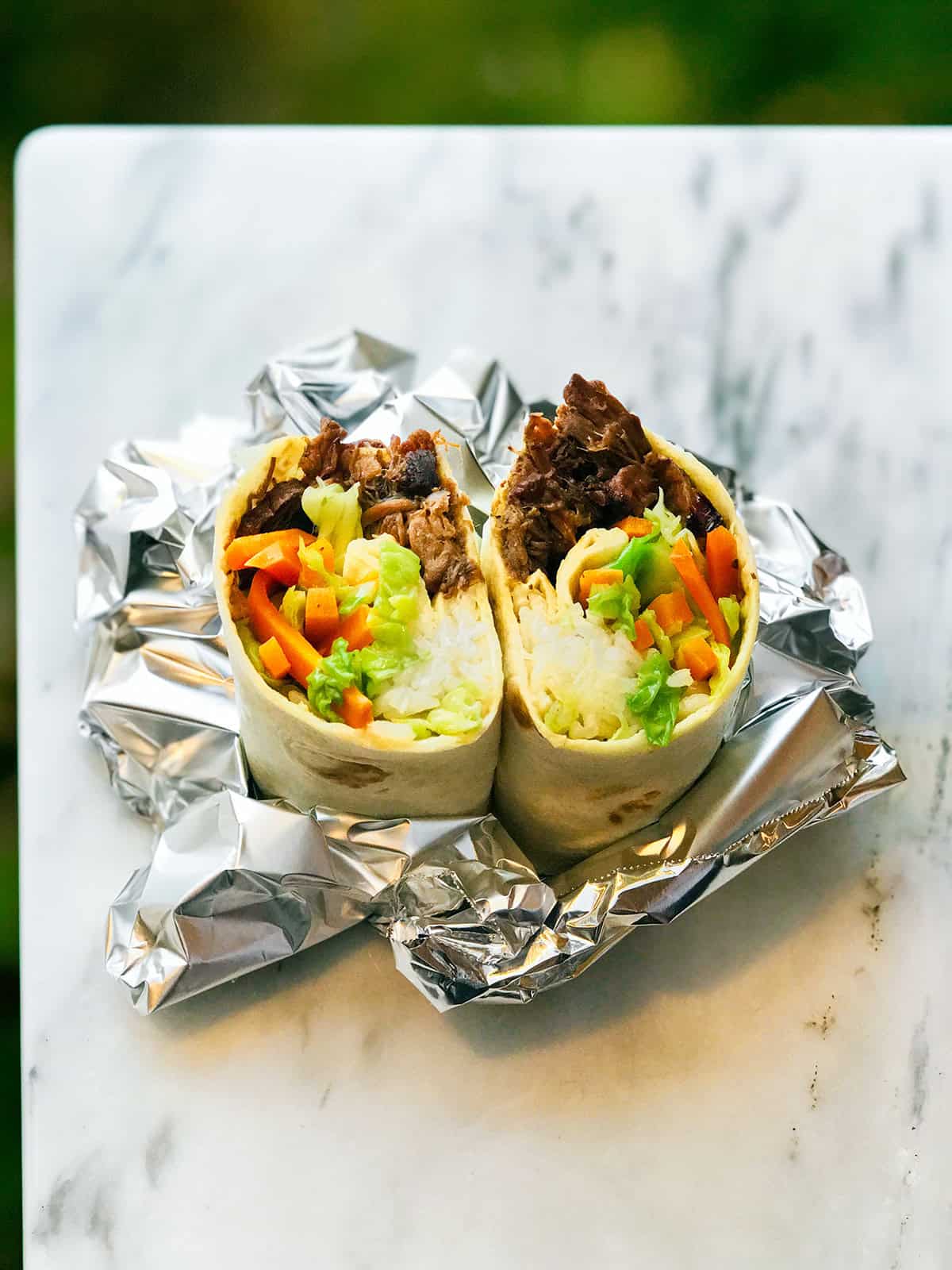 A burrito cut in half on a sheet of aluminum foil, showing rice, bulgogi shredded beef, lettuce, and carrot sticks inside, placed on a marble surface.