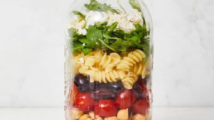 A glass jar filled with layered salad ingredients including chickpeas, tomatoes, pasta, black beans, greens, and feta cheese sits beside a metal lid—perfect for those looking for Healthy Lunch Ideas.