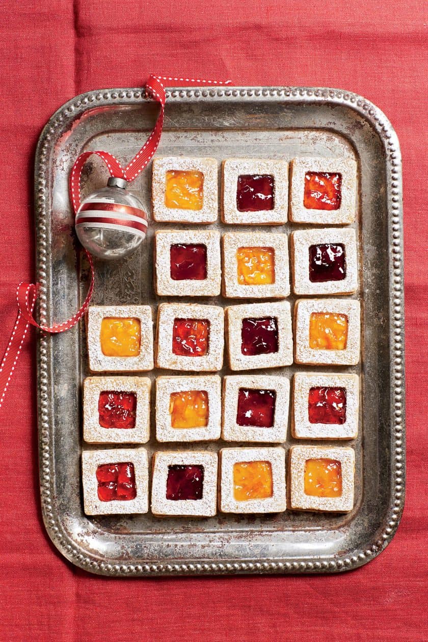 A silver tray with five rows of Vintage Christmas Cookies-square treats filled with red and yellow jam, dusted with powdered sugar-sits beside a silver ornament with a red ribbon on a festive red tablecloth.