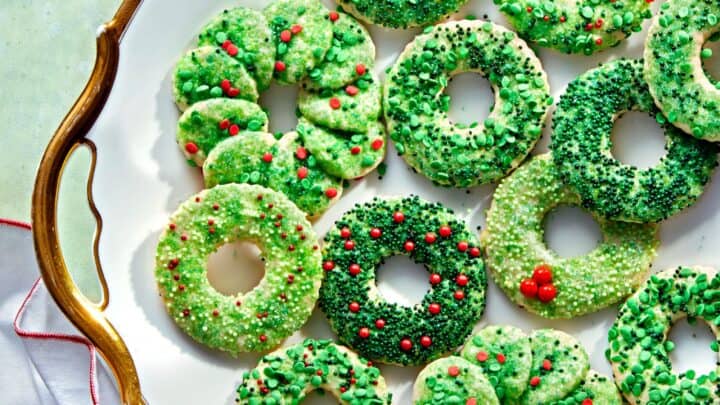 A white plate with twelve green holiday wreath cookies, decorated with red, green, and white sprinkles, sits on a green surface with a white napkin nearby—perfect Vintage Christmas Cookies for festive gatherings.