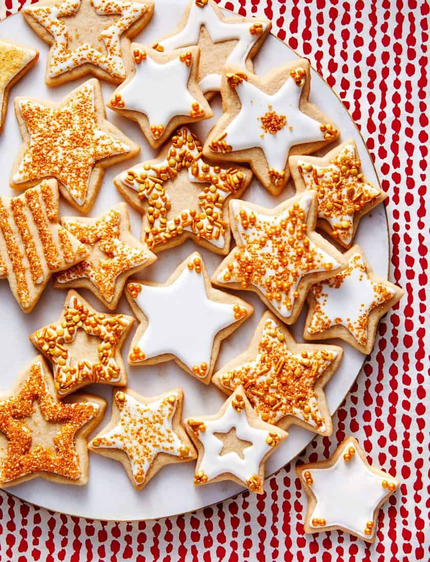 A plate of star-shaped Vintage Christmas Cookies, decorated with white icing and gold sprinkles, arranged on a red and white patterned surface.