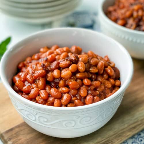 A white bowl filled with Instant Pot Baked Beans in a rich sauce sits on a wooden board, with another bowl of beans and a sprig of herbs blurred in the background.