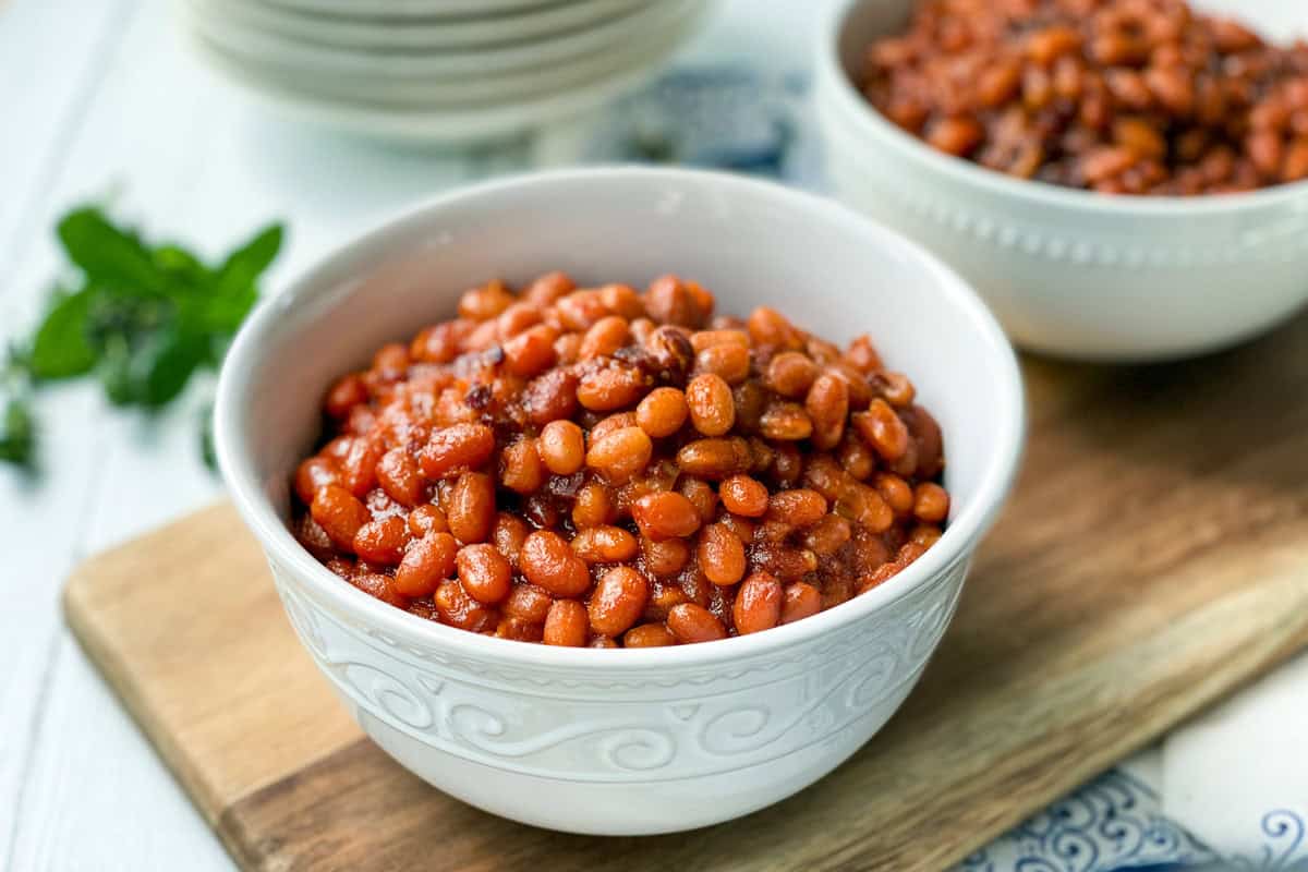 A white bowl filled with Instant Pot Baked Beans in a rich sauce sits on a wooden board, with another bowl of beans and a sprig of herbs blurred in the background.