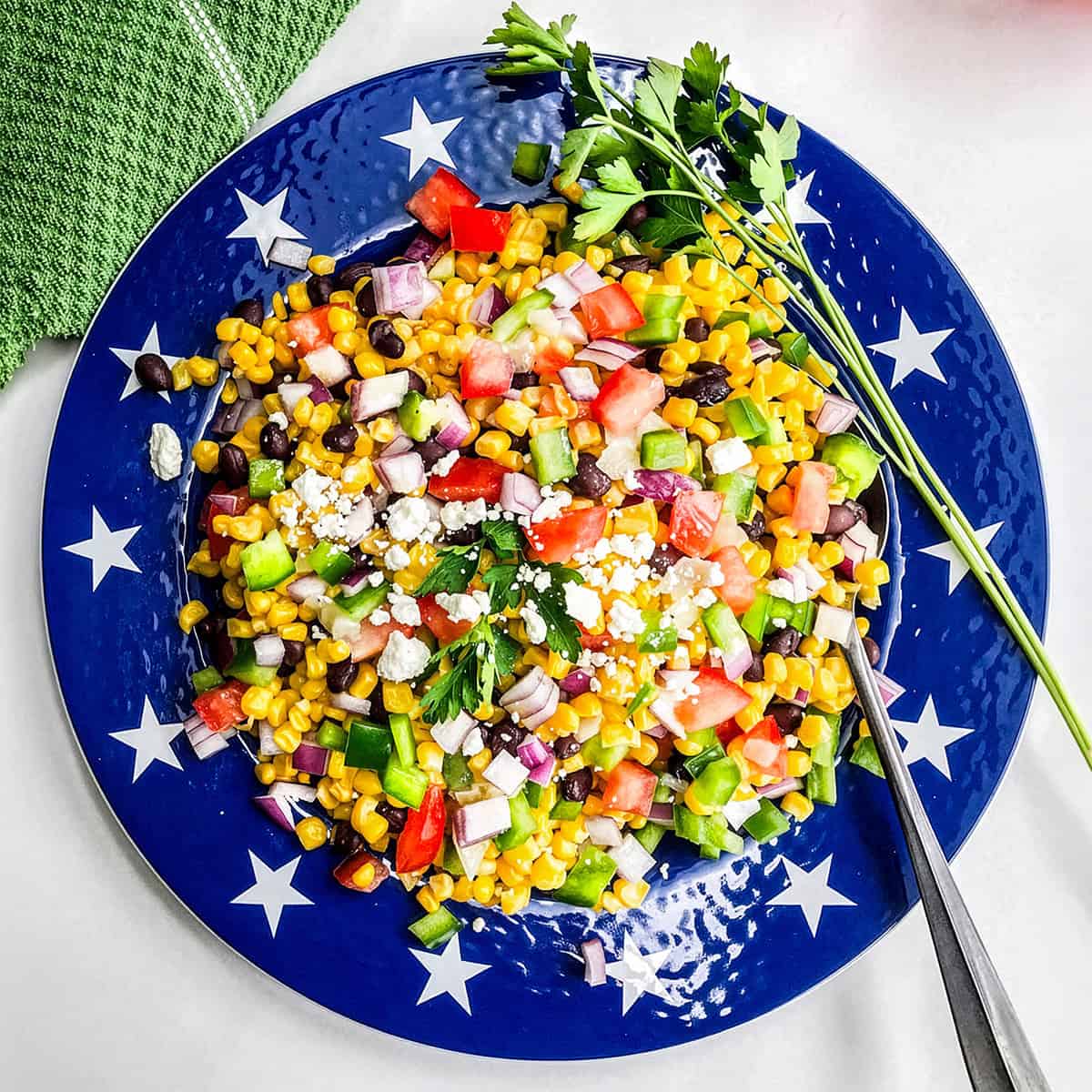 A blue plate with white stars holds a vibrant salad of corn, black beans, tomatoes, red onion, green peppers, feta cheese, and parsley-an enticing addition to your Summer Side Dishes lineup, complete with serving utensils and fresh herbs on the side.