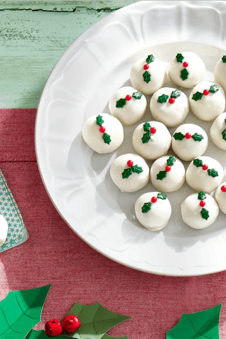 A white plate with round white Christmas candy truffles, decorated with green holly leaves and red berries, sits on a red placemat.