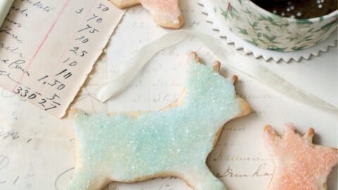Iced animal-shaped sugar cookies and round cookies with white sprinkles are arranged on handwritten recipe papers beside a pencil and a decorative tin, creating a nostalgic scene of vintage Christmas cookies.
